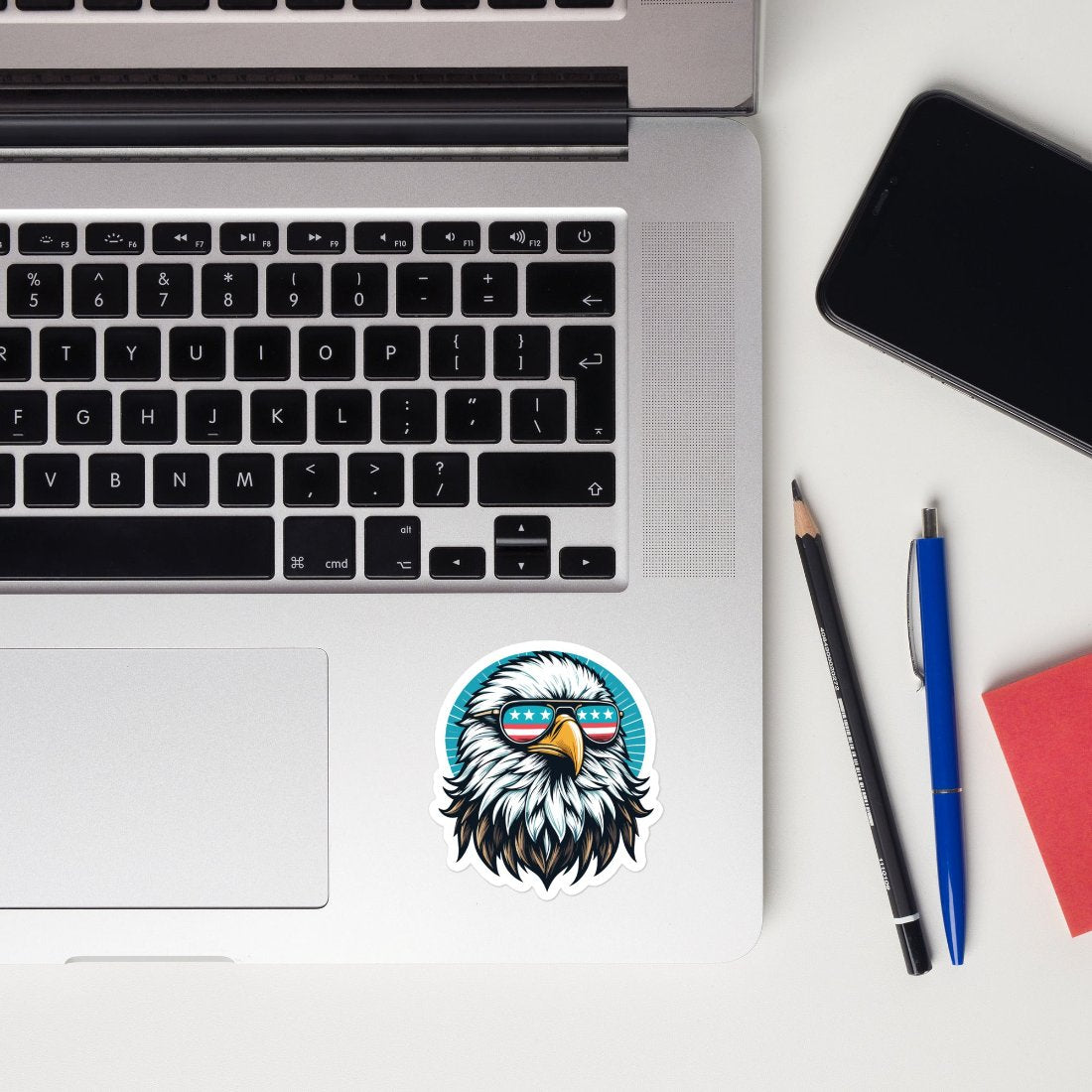 A patriotic bald eagle sticker placed on the bottom-right corner of a laptop. The eagle is wearing sunglasses with the American flag design on the lenses. The setup includes a pencil, pen, notebook, and smartphone on a desk, creating a workspace vibe with a touch of American pride.